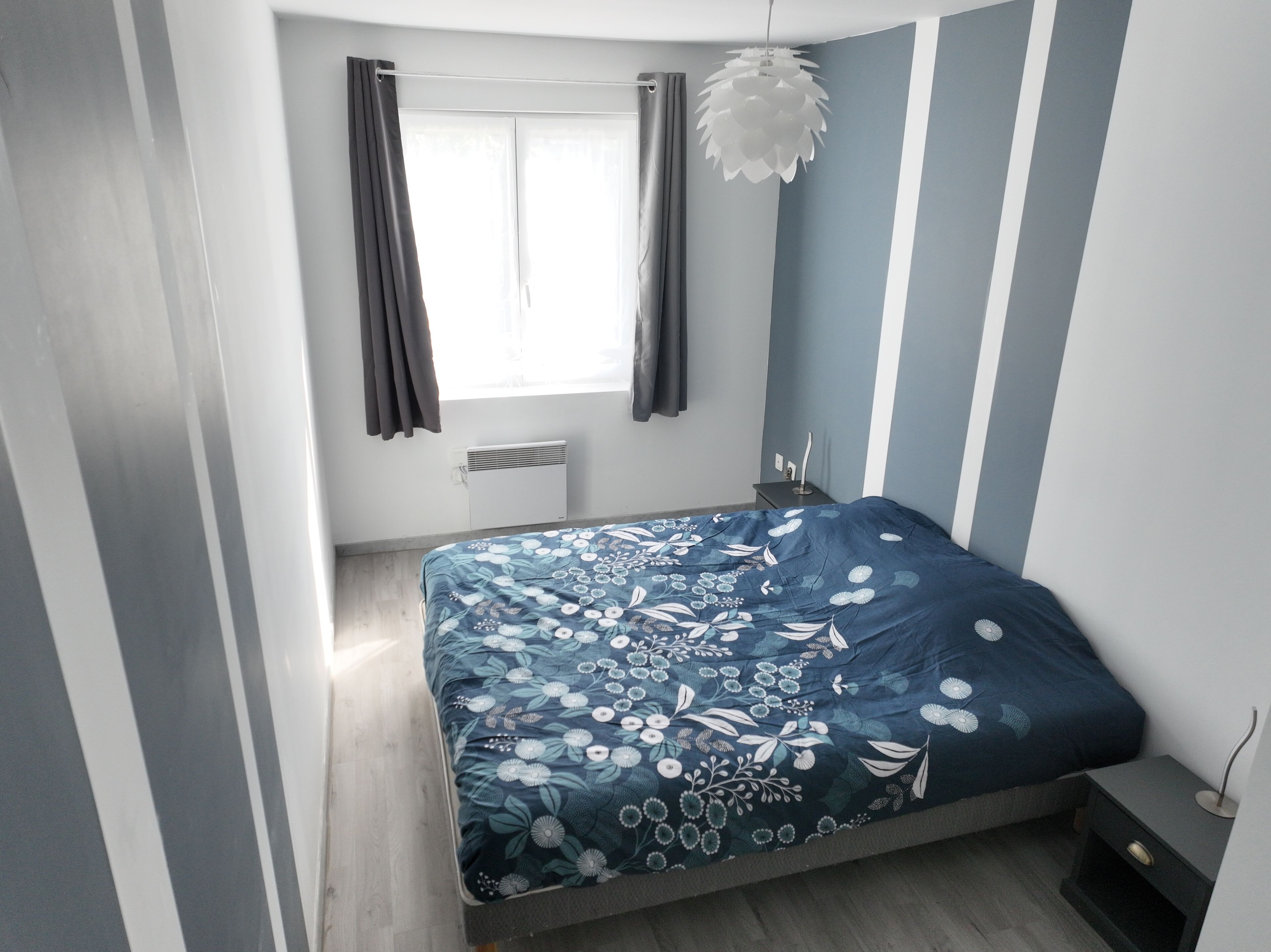 A bedroom with a bed, a window with curtains, a radiator, a bedside table, and a hanging light fixture.