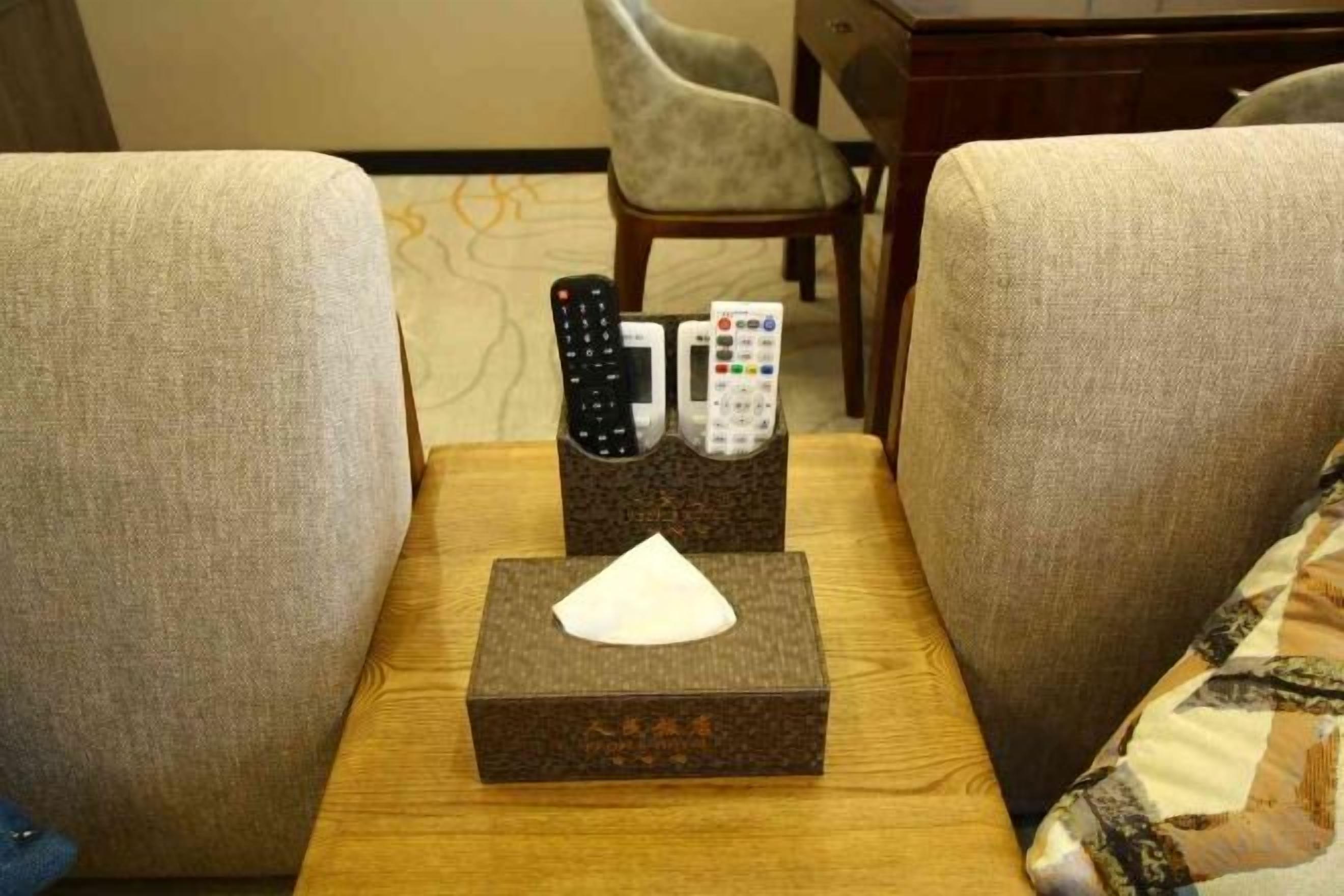 Uma mesa lateral de madeira com controles remotos, um estojo de lenços de papel e um almofada entre dois sofás.