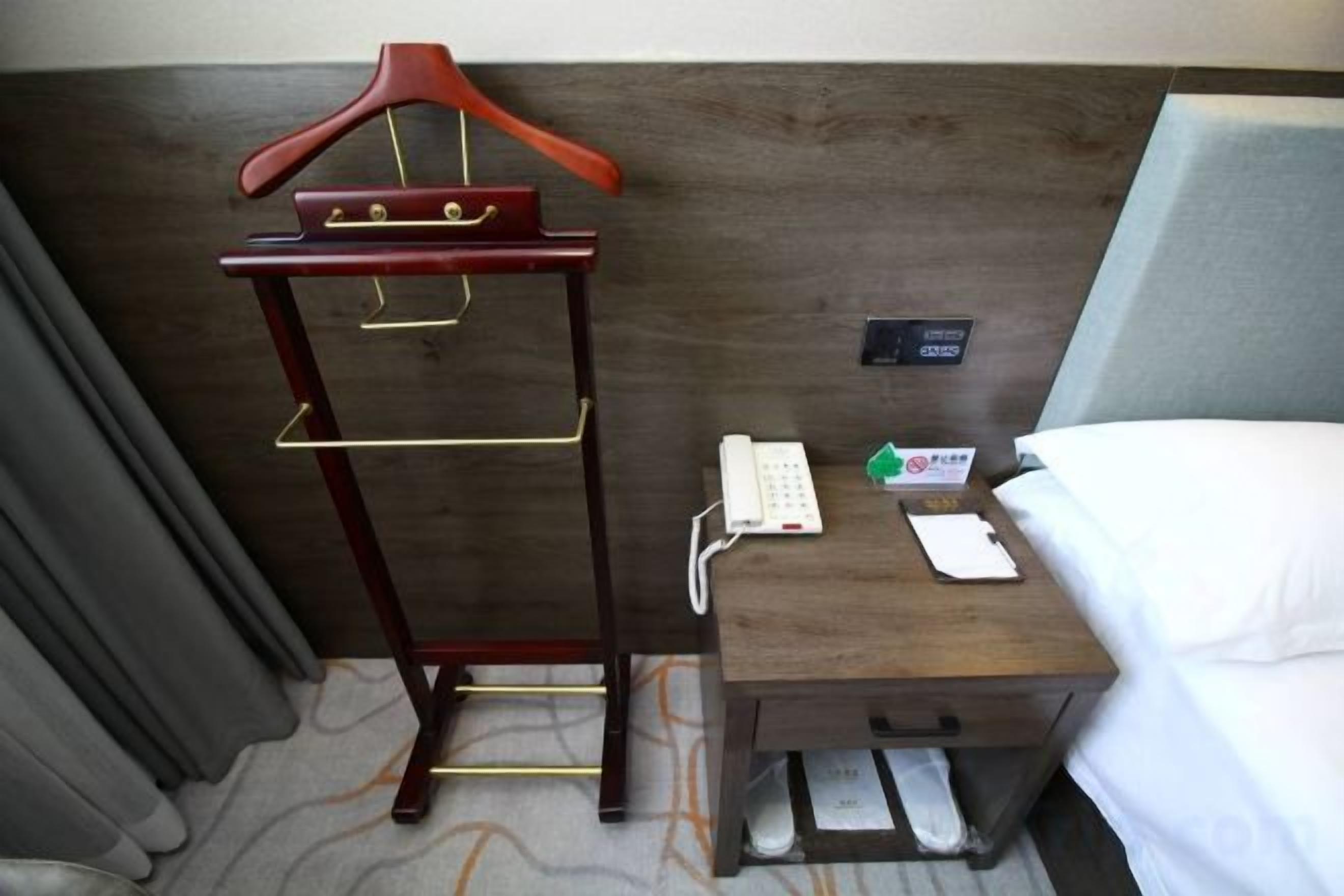 Una habitación de hotel con una mesita de noche de madera, un teléfono, un bloc de notas y un perchero.