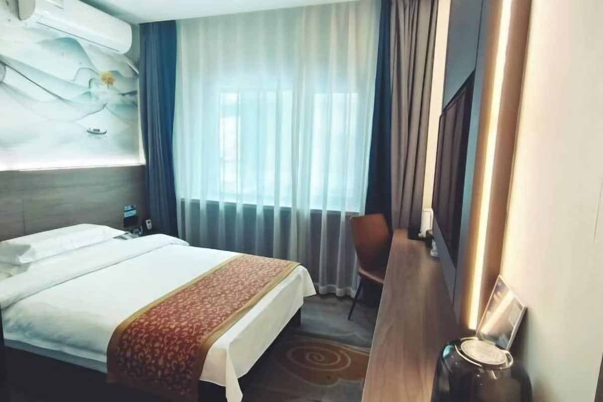 Quarto de hotel com cama, escrivaninha, cadeira, janela com cortinas e ar condicionado fixado na parede.