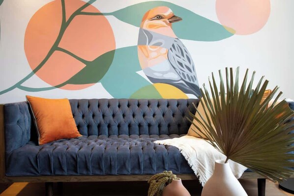 A dark blue tufted sofa with an orange pillow, a white vase with a plant, and a colorful abstract wall art.