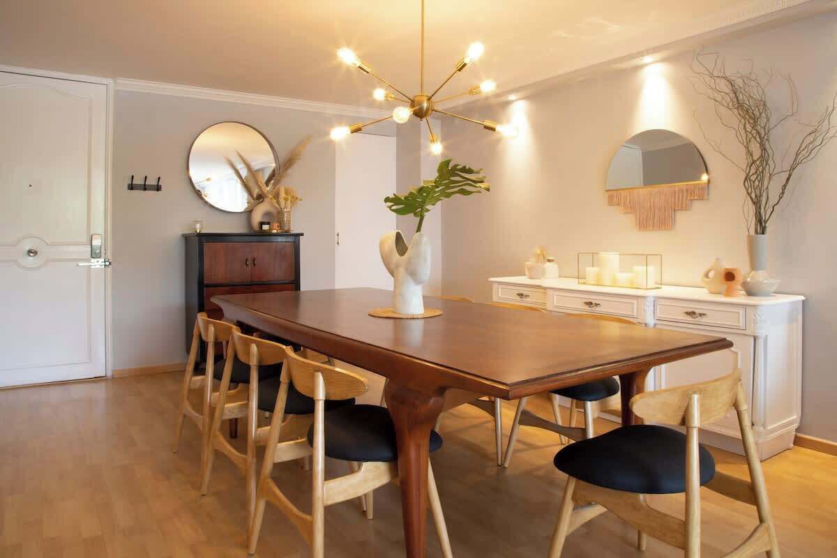 Zona pranzo con tavolo in legno, sei sedie, un vaso con una pianta, uno specchio e un lampadario moderno.