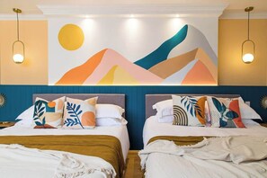 A hotel room with two beds, each with a headboard and multiple pillows. There is a large abstract painting above the beds and two pendant lights hanging from the ceiling.