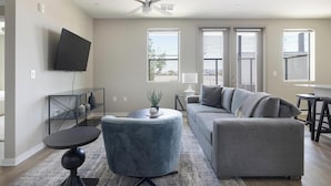 Ein modernes Wohnzimmer mit einem grauen Sofa, einem runden Ottomanen, einem an der Wand montierten Fernseher und einem Deckenventilator.