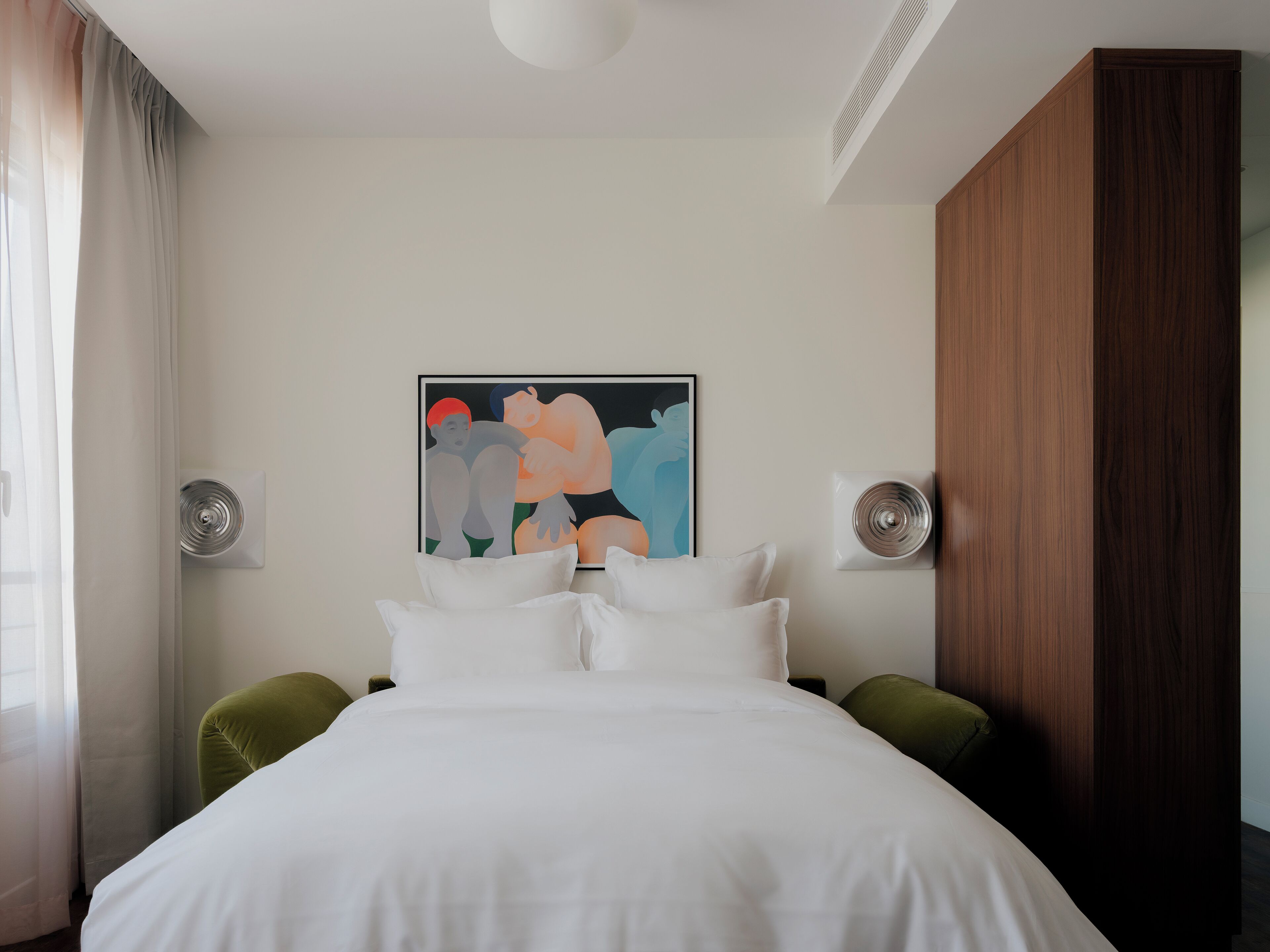 A modern hotel room with a large bed, green upholstered headboard, white bedding, a wall-mounted air conditioning unit, and a framed abstract painting.