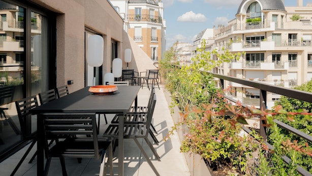 A balcony with a table and chairs, surrounded by greenery and overlooking a row of buildings with balconies.
