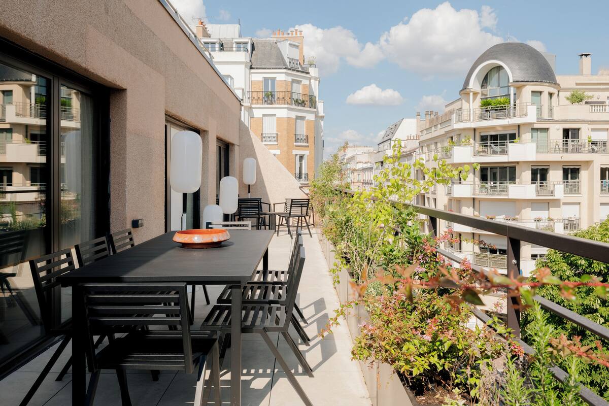 Een balkon met een tafel en stoelen, omgeven door groen en met uitzicht op een rij gebouwen met balkons.