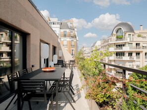 Ein Balkon mit Tisch und Stühlen, umgeben von Grünpflanzen und mit Blick auf eine Reihe von Gebäuden mit Balkonen.