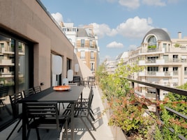 A balcony with a table and chairs, surrounded by greenery and overlooking a row of buildings with balconies.