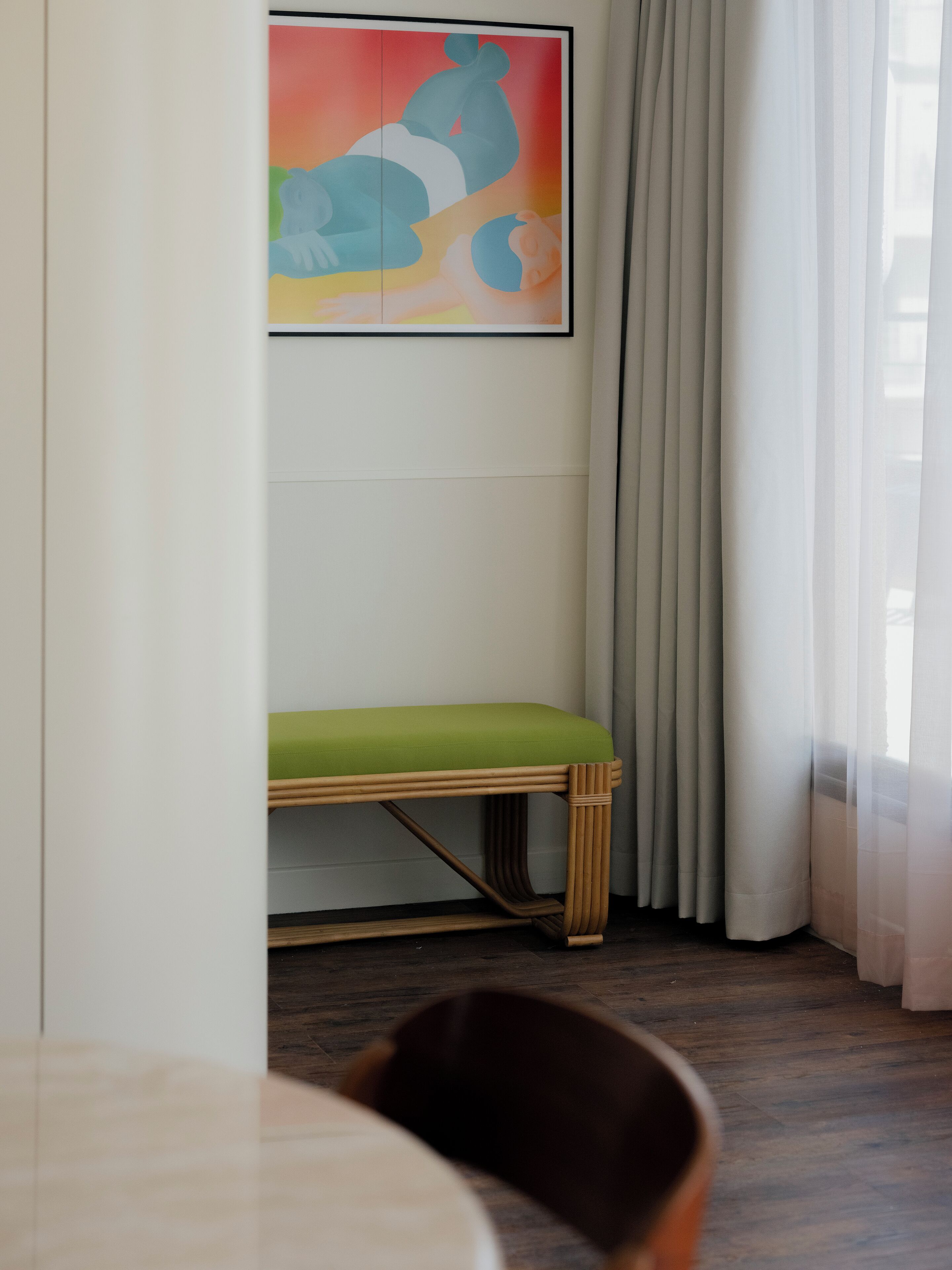 A room with a wooden bench, a framed abstract painting, and a window with sheer curtains.