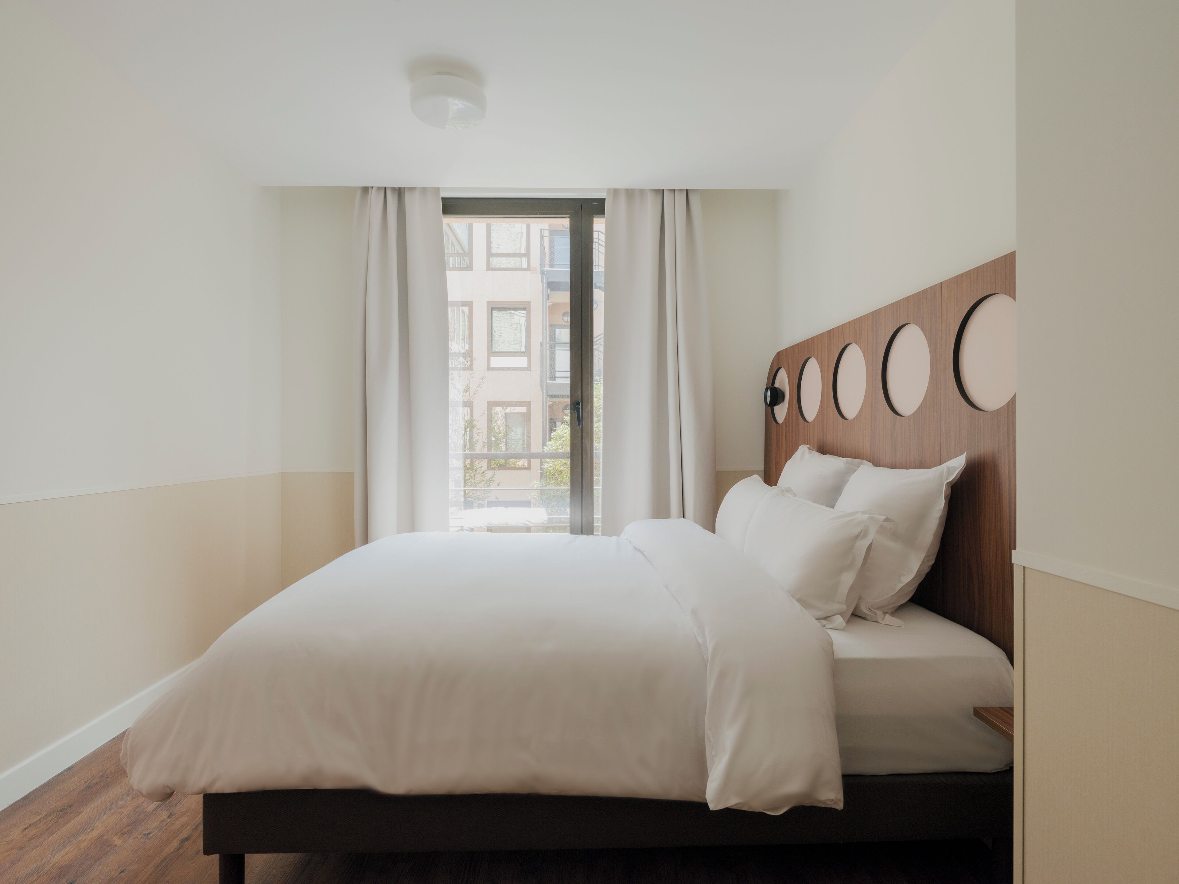 Quarto de hotel moderno com uma cama grande, cabeceira de madeira com detalhes circulares e vista para prédios pela janela.