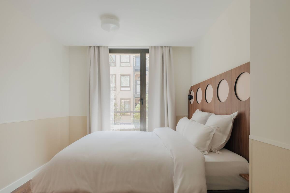 Een moderne hotelkamer met een groot bed, een houten hoofdeinde met ronde decoraties en een uitzicht op gebouwen door een raam.