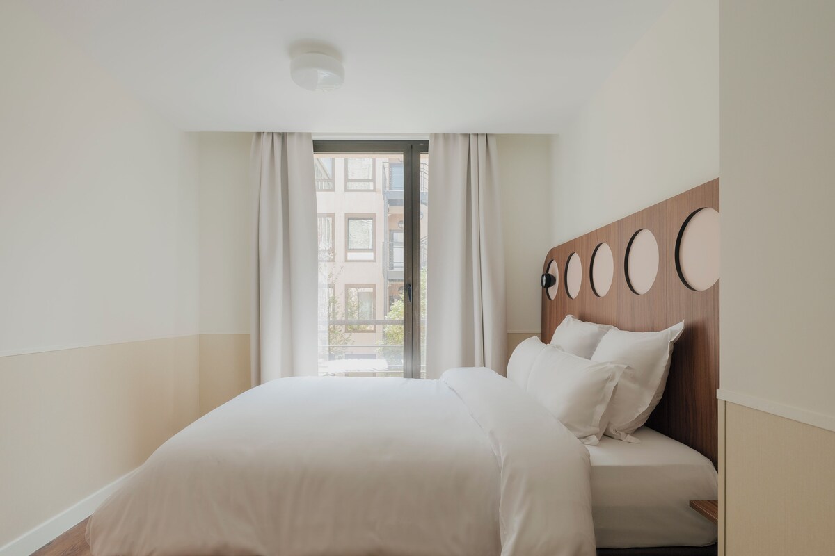 Een moderne hotelkamer met een groot bed, een houten hoofdeinde met ronde decoraties en een uitzicht op gebouwen door een raam.