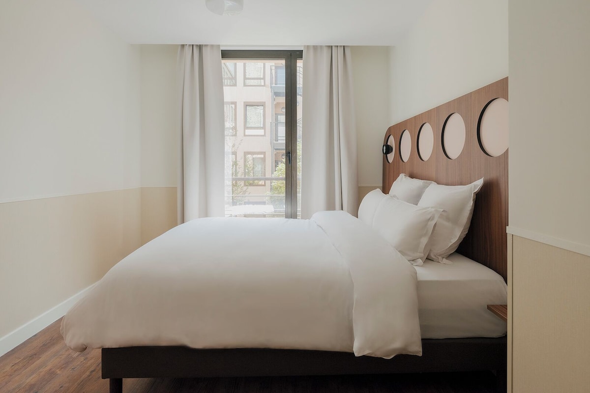 A modern hotel room with a large bed, a wooden headboard with circular decorations, and a view of buildings through the window.