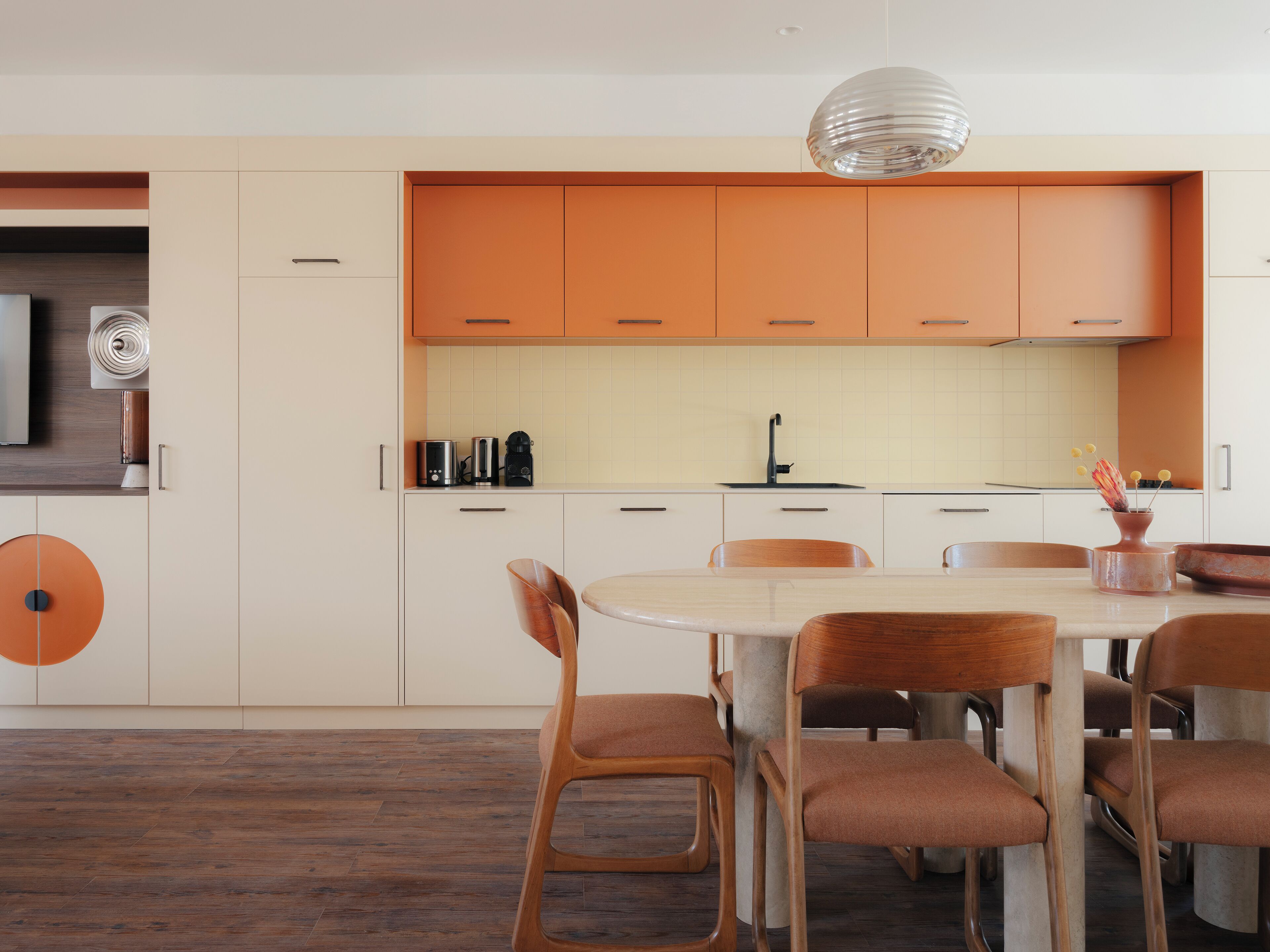 A modern kitchen with orange cabinets, a white countertop, and a dining area with wooden chairs.