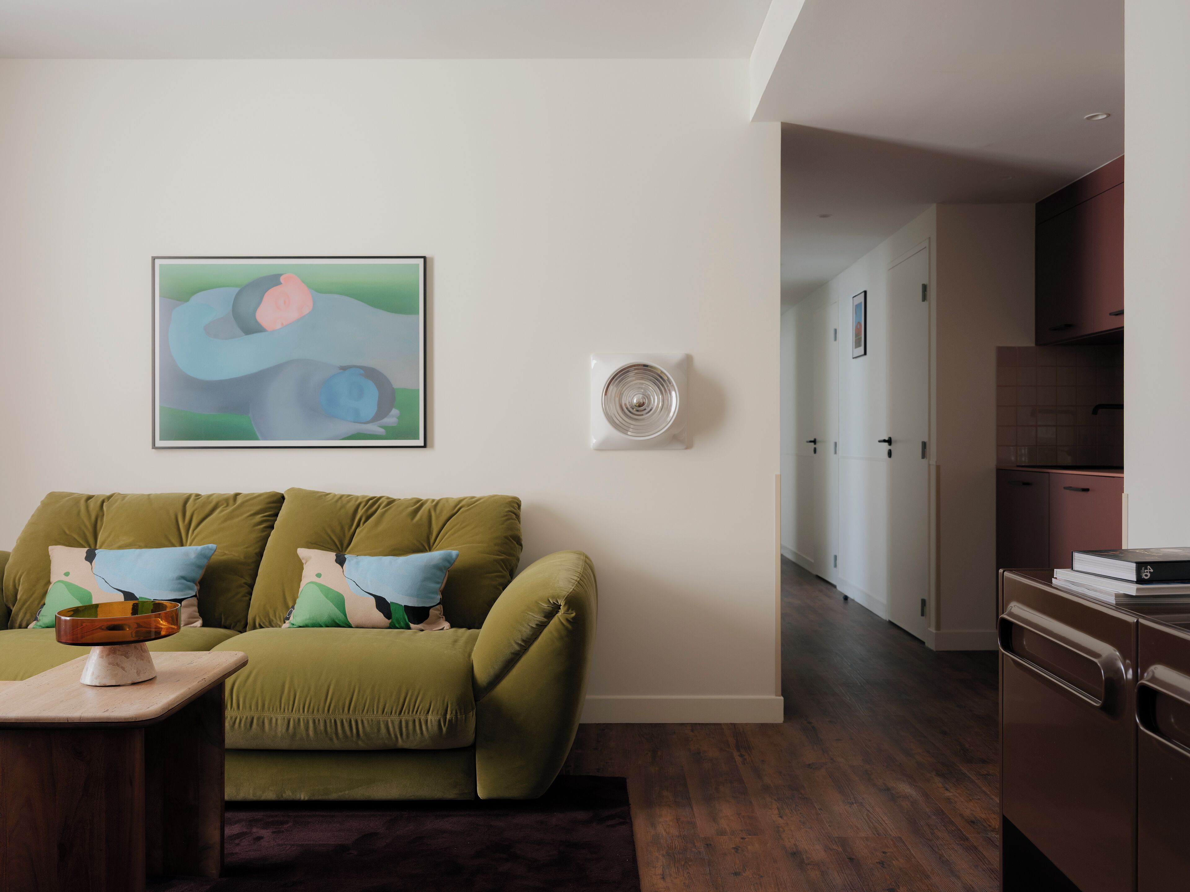 A modern living room with a green sofa, a coffee table, and a wall-mounted fan. A kitchen is visible in the background.
