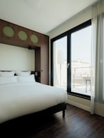 A hotel room with a large bed, wooden headboard, and a balcony with a view of buildings.