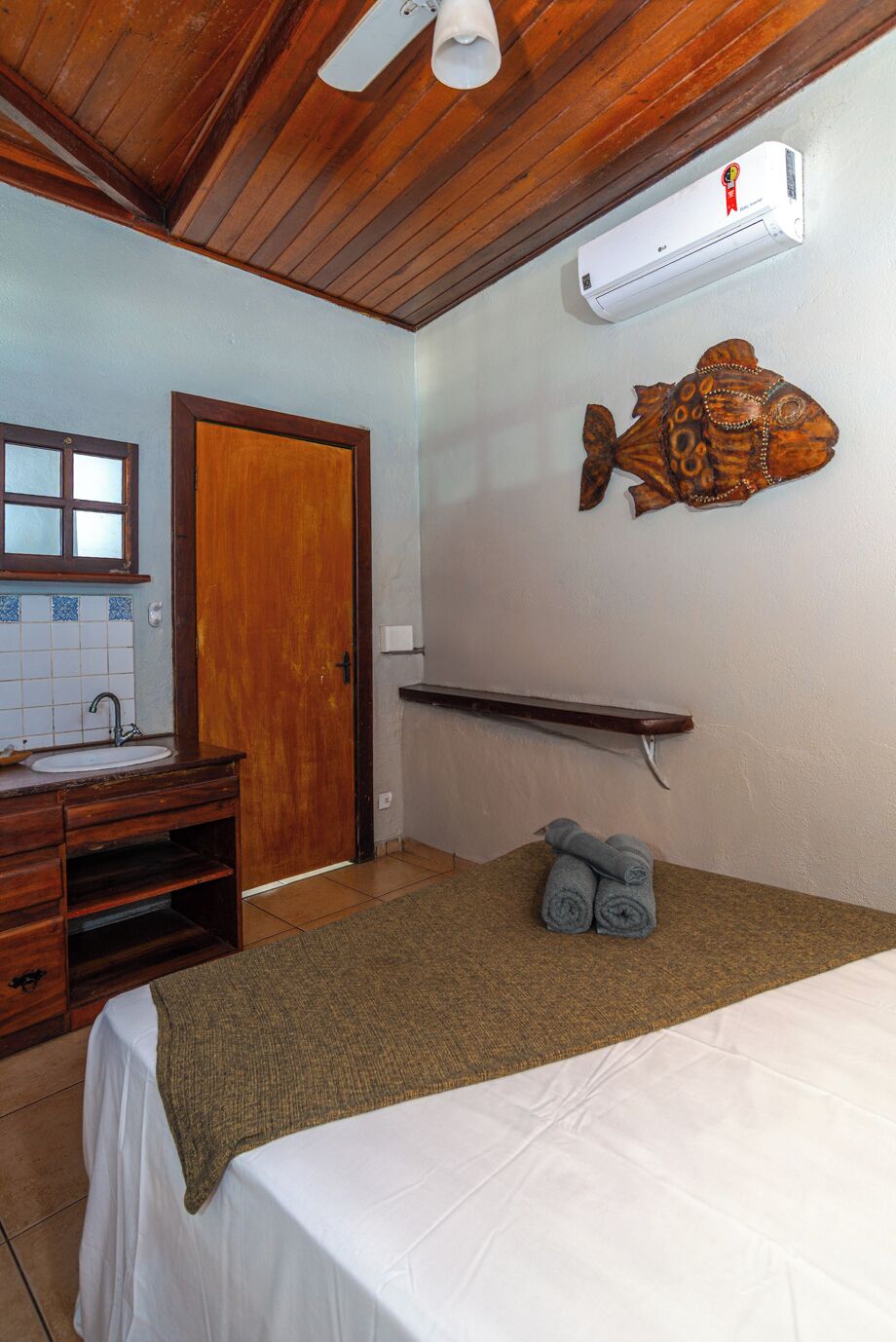 A room with a wooden ceiling, a white bed, a wooden vanity with a sink, a towel rack, and a wall-mounted air conditioner.