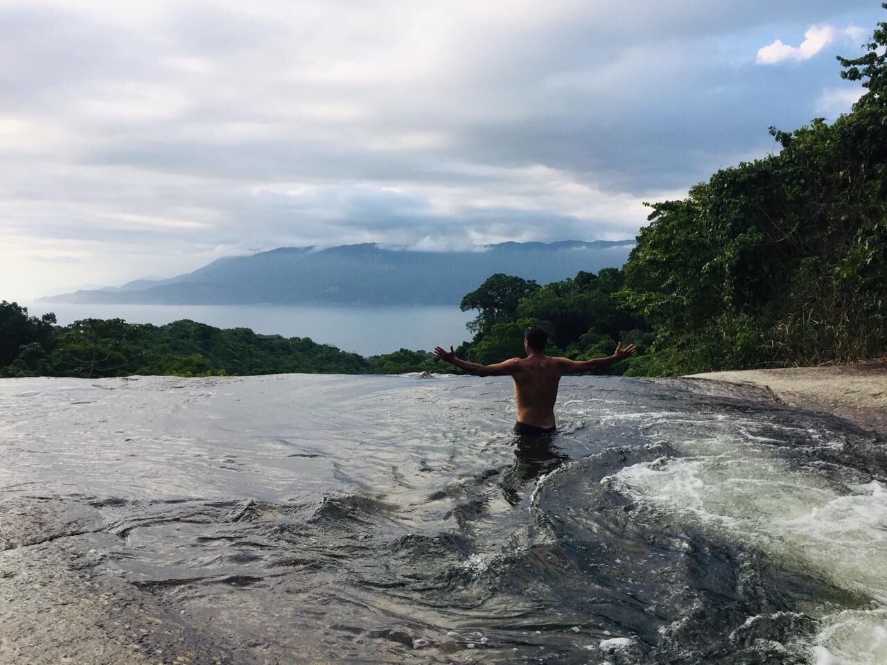 Una persona de pie en un río con los brazos extendidos, rodeada de follaje y un cielo nublado.