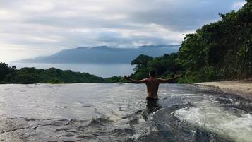 Una persona de pie en un río con los brazos extendidos, rodeada de follaje y un cielo nublado.