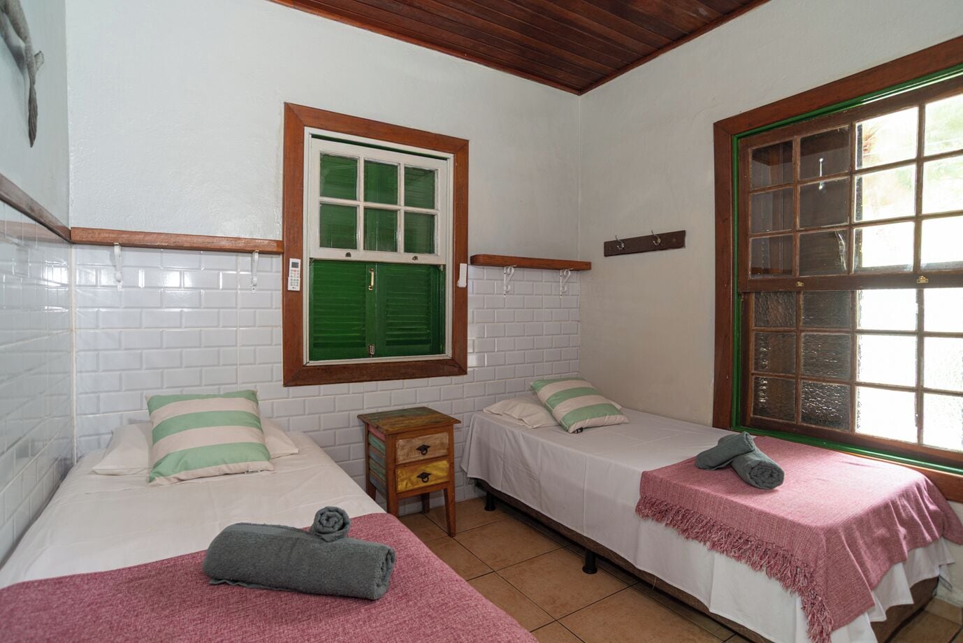 A room with two beds, a small wooden nightstand, a window with green shutters, and a tiled wall.