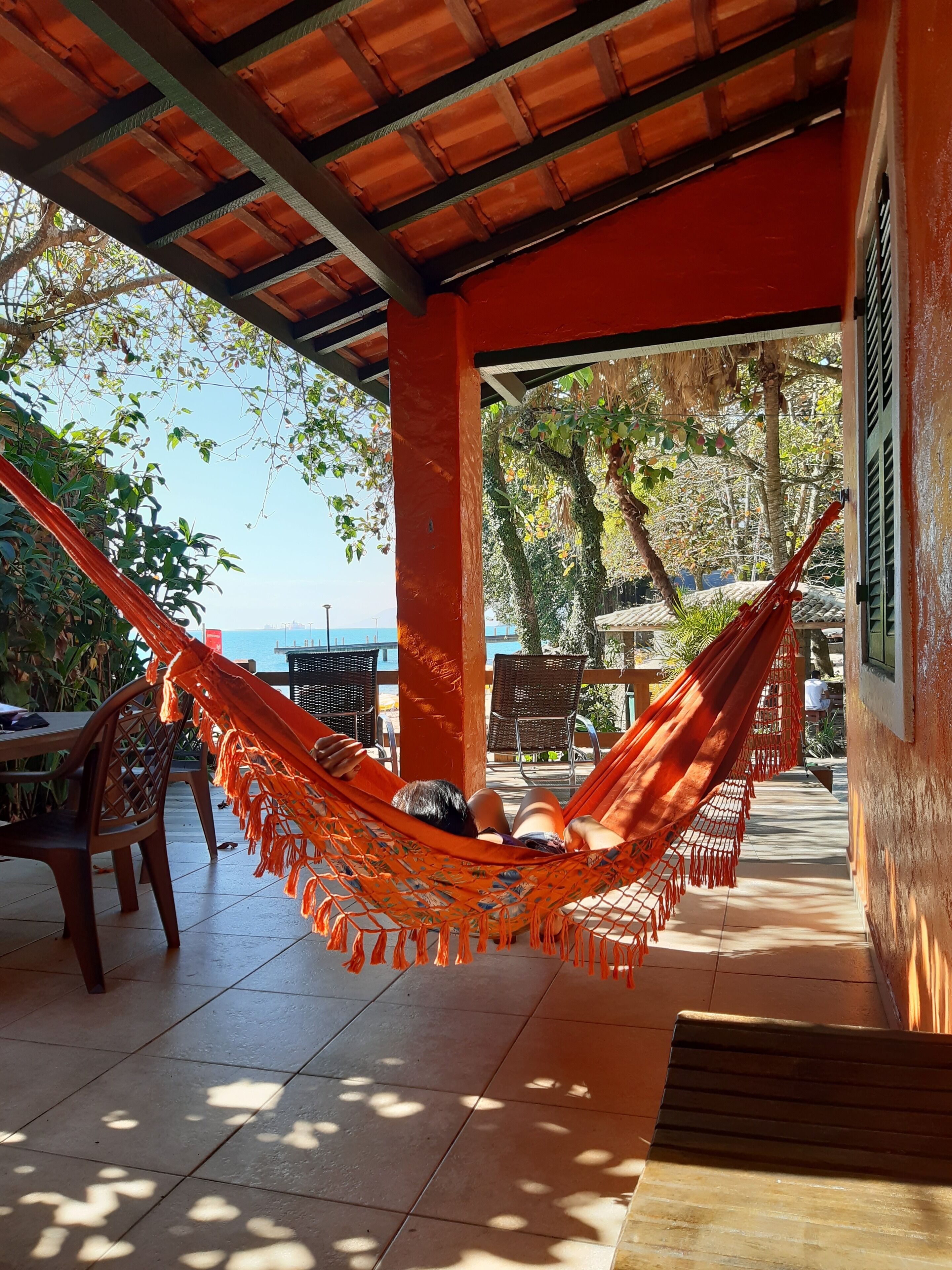 A covered outdoor seating area with a hammock, chairs, and a view of the sea.