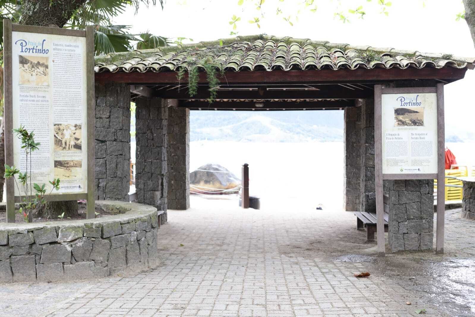 A stone archway leading to a beach with a signboard and a wooden pergola overhead.