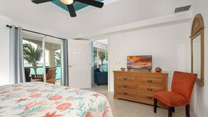 Un dormitorio con cama, cómoda de madera, silla, televisor y vista al océano a través de una puerta corrediza.