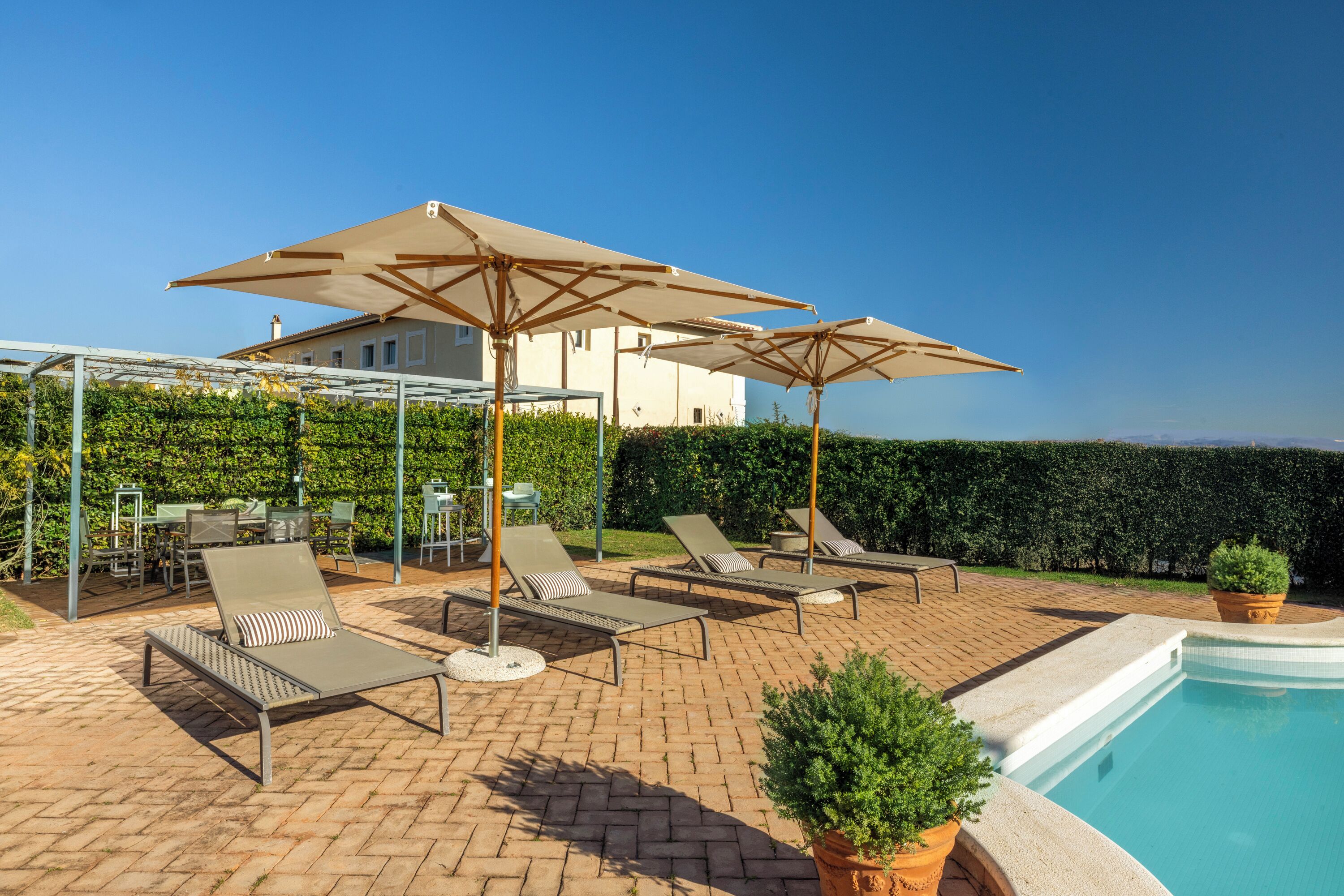 Un área junto a la piscina con sillones de descanso y sombrillas, un patio de ladrillo y vista a un edificio al fondo.