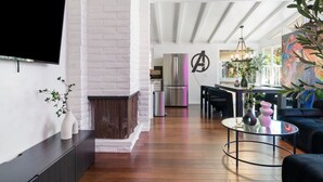 A modern living room with a white brick fireplace, a flat-screen TV mounted on the wall, a dining area with a kitchen in the background, and a large painting on the wall.
