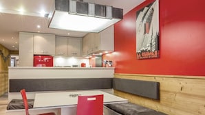 A modern dining area with a white table and red chairs, a kitchen in the background, and a red wall with a poster.