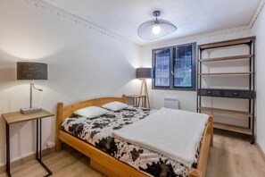 A bedroom with a wooden bed, a bedside table, a lamp, a radiator, and a window with blinds.