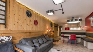A cozy interior with a sofa, chairs, a wooden wall, a clock, and a kitchen area.