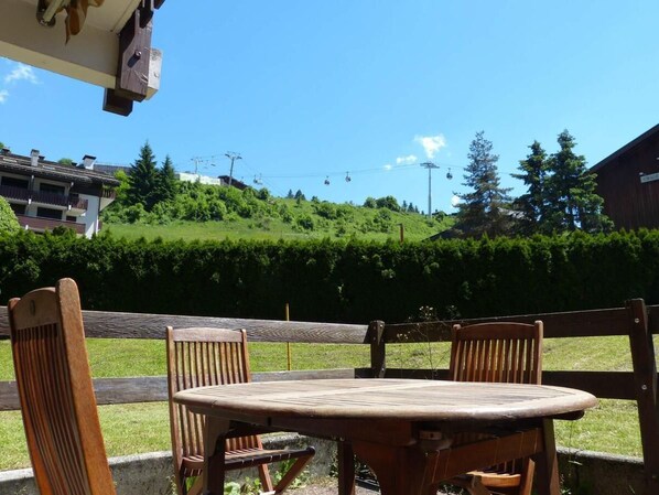 Une table et des chaises en bois sont placées à l’extérieur, offrant une vue sur une colline verdoyante et un bâtiment en arrière-plan.