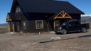 Une grande maison en bois sombre, surmontée d’un toit en métal, avec un camion garé devant et un paysage montagneux en arrière-plan.