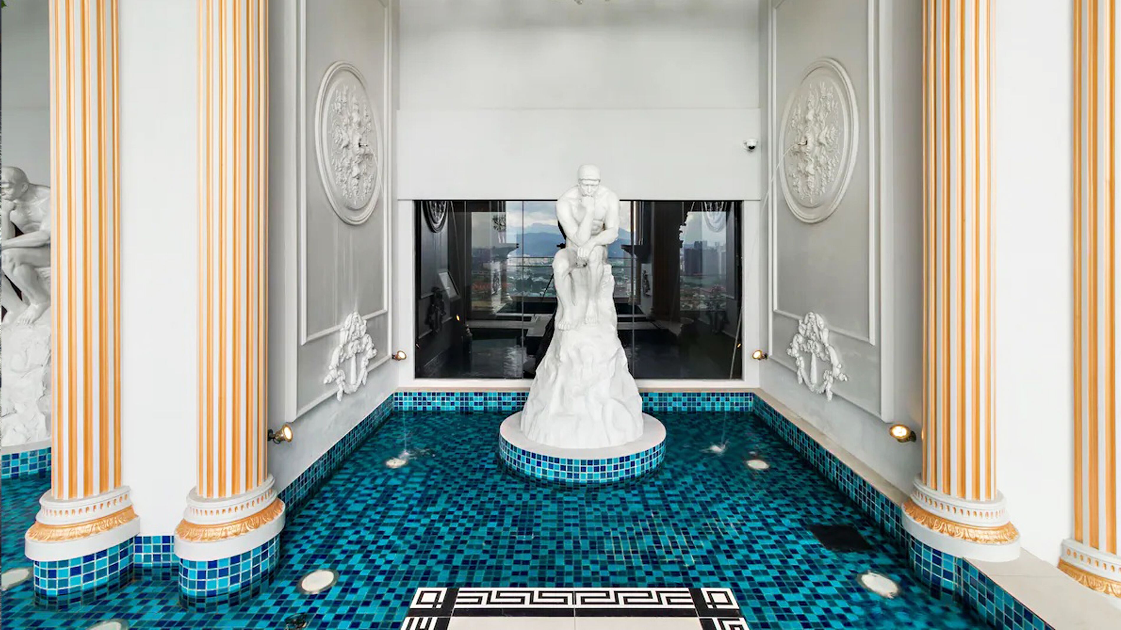 A luxurious indoor pool area with a central statue, ornate columns, and a tiled floor.