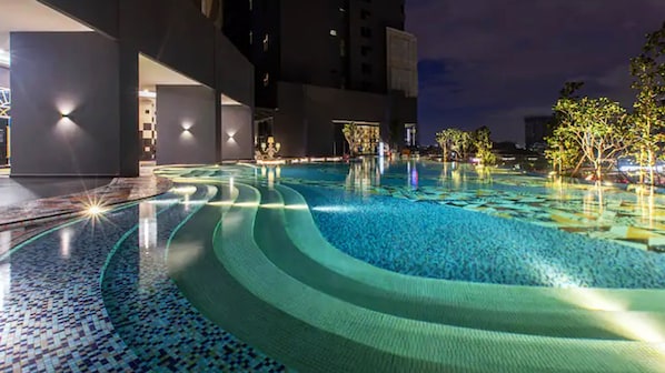 Une piscine nocturne au design ondulant, entourée d’un bâtiment moderne aux murs illuminés, offrant une vue dégagée sur le ciel.