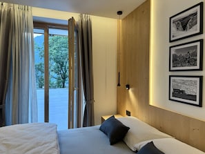 A bedroom with a bed, two pillows, a nightstand, and a wall-mounted lamp. There are curtains on the window, and three framed pictures on the wall.