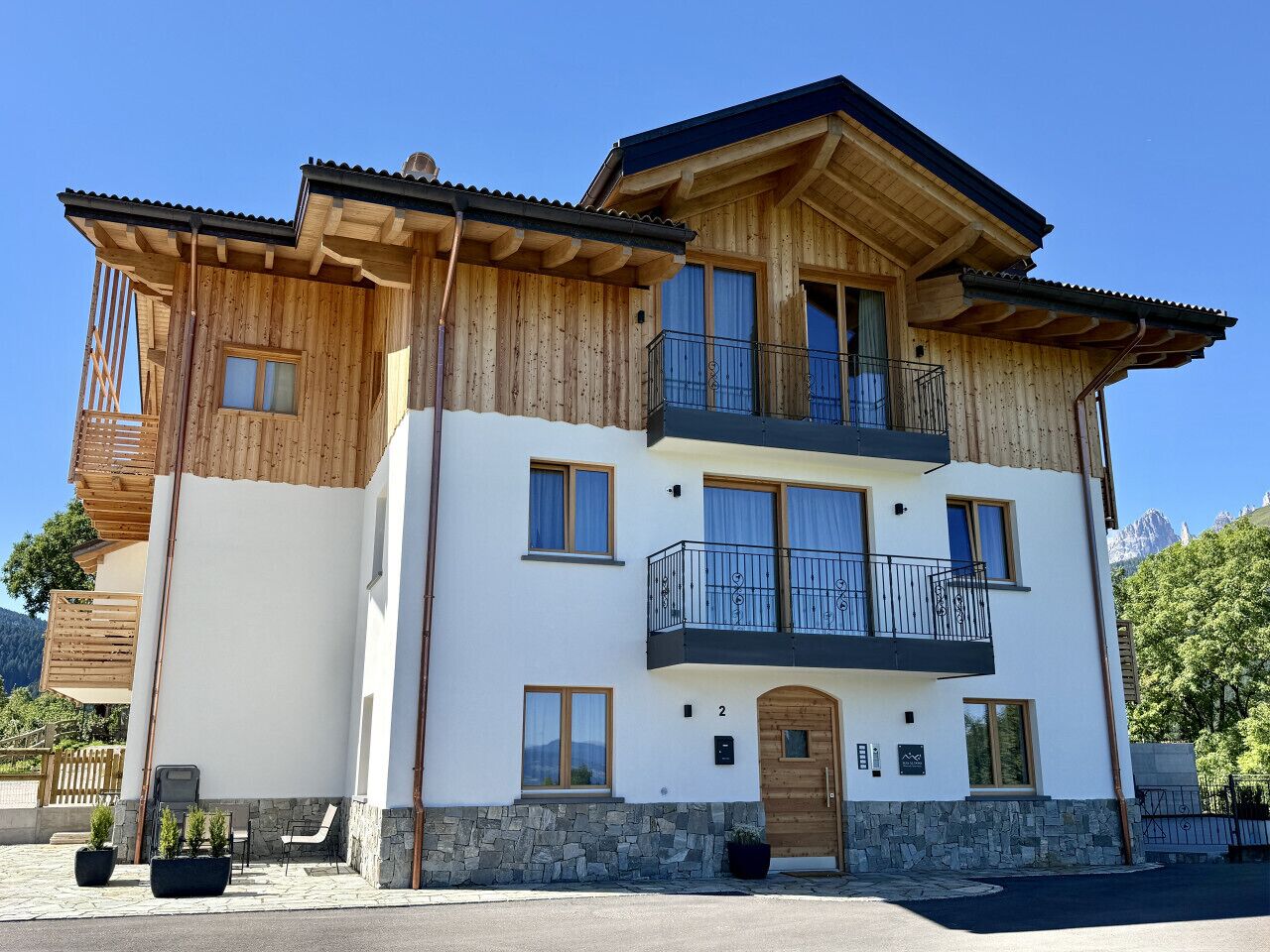 A two-story wooden house with a balcony, stone foundation, and a mountain backdrop.