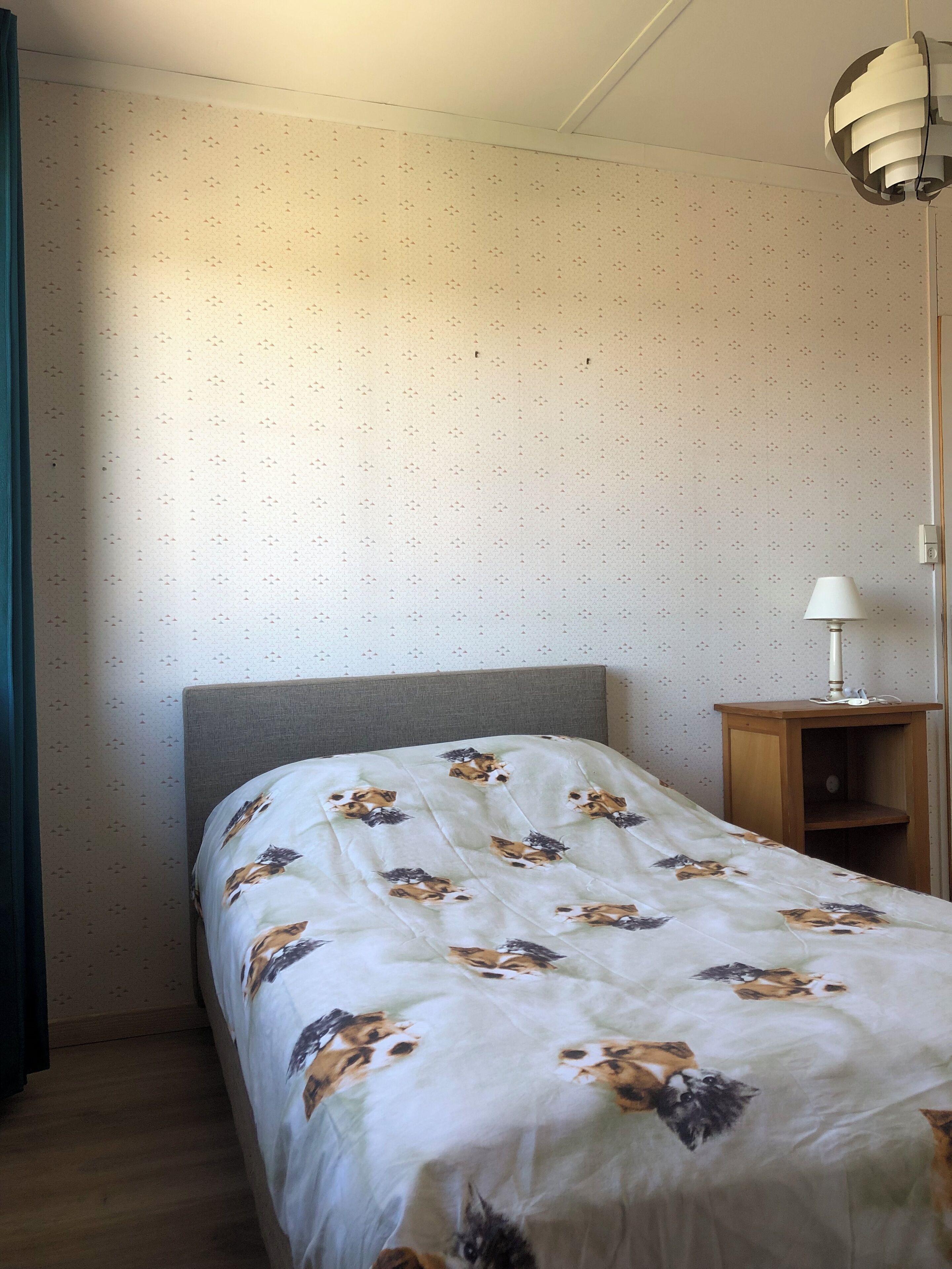 En pænt redt seng med et blomstret sengetæppe, et natbord med en lampe og en tapetvæg.