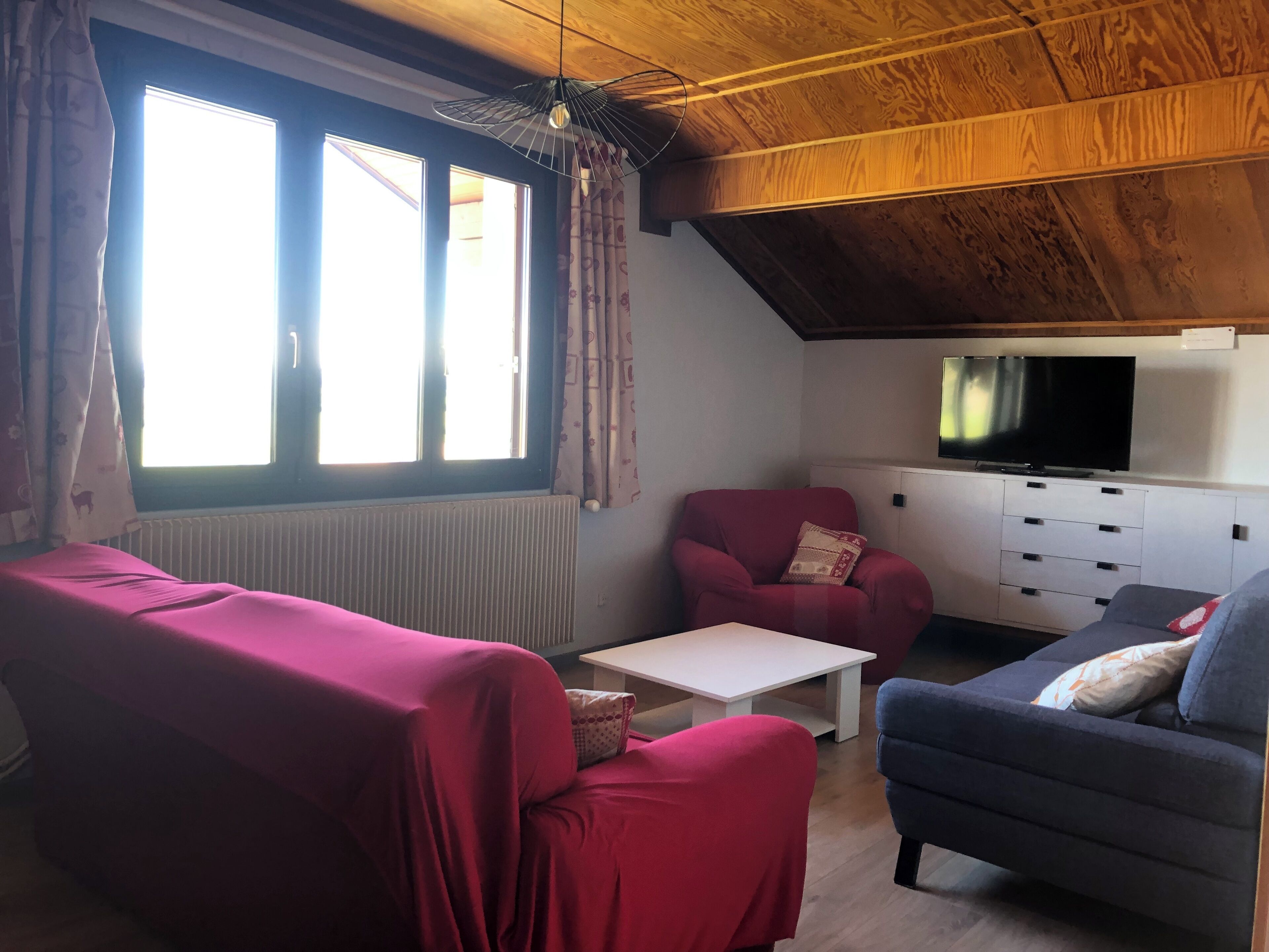 Una sala de estar acogedora con un sofá rojo, un sillón azul y una mesa de centro blanca. Hay un televisor sobre un mueble blanco, y la habitación tiene vigas de madera en el techo.