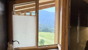A room with a large window, a wooden ceiling, and a small shelf with a decorative item. - Attico Stella Alpina (Andalo)