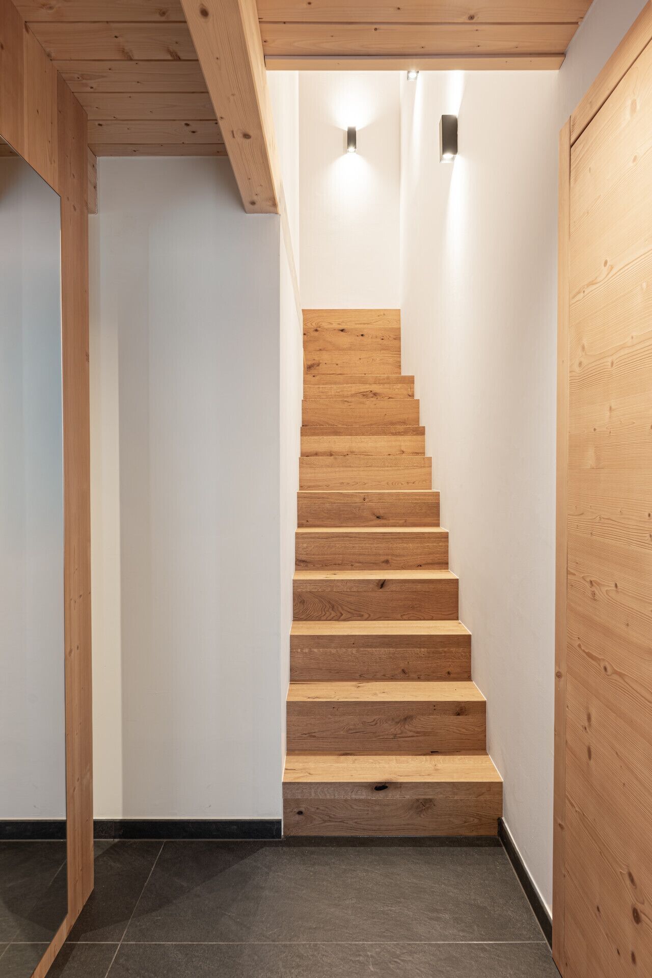 A wooden staircase in a hallway with white walls and a dark floor.