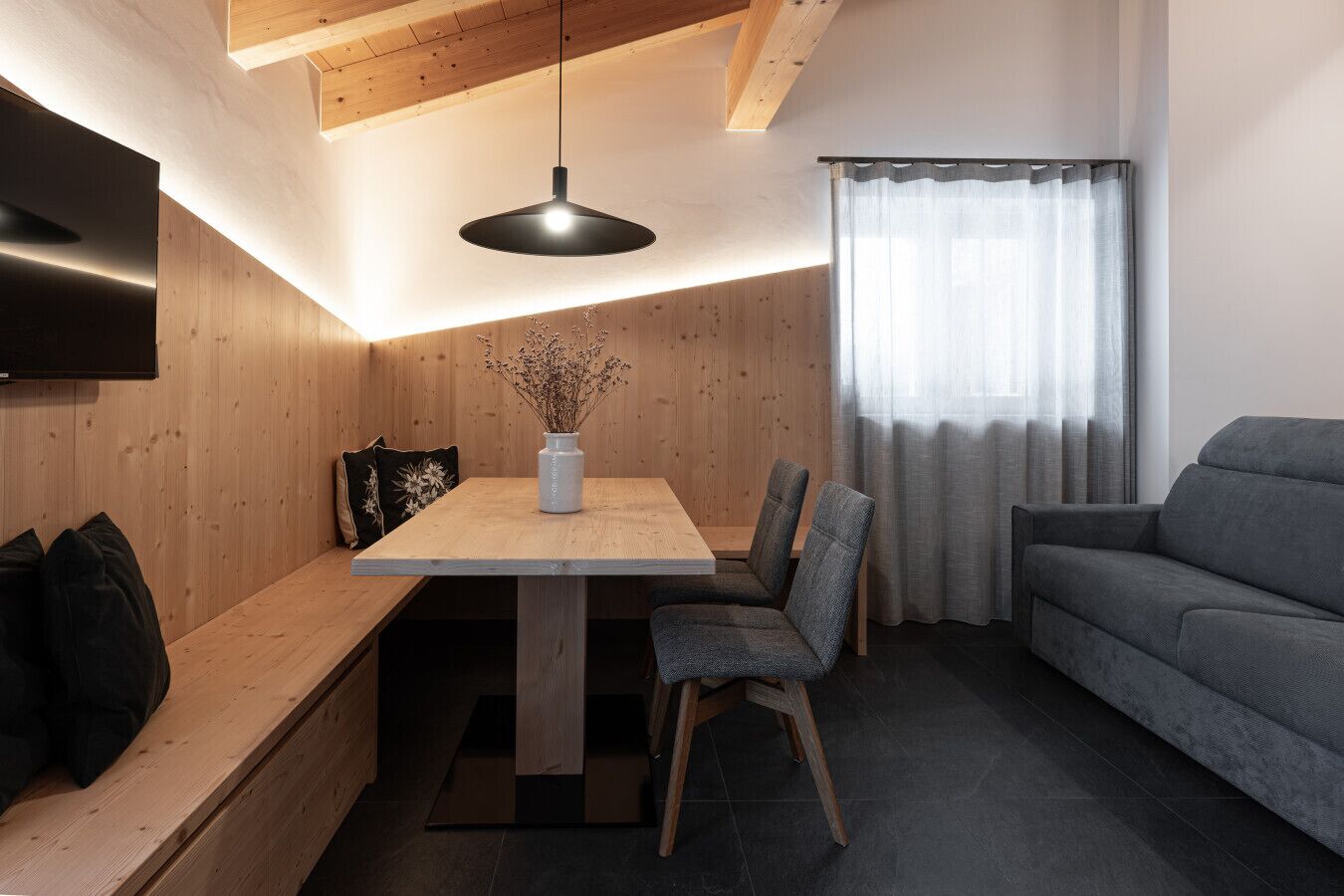 Ein modernes Wohnzimmer mit einem Holzesstisch, zwei Stühlen, einem grauen Sofa und einem an der Wand montierten Flachbildfernseher.