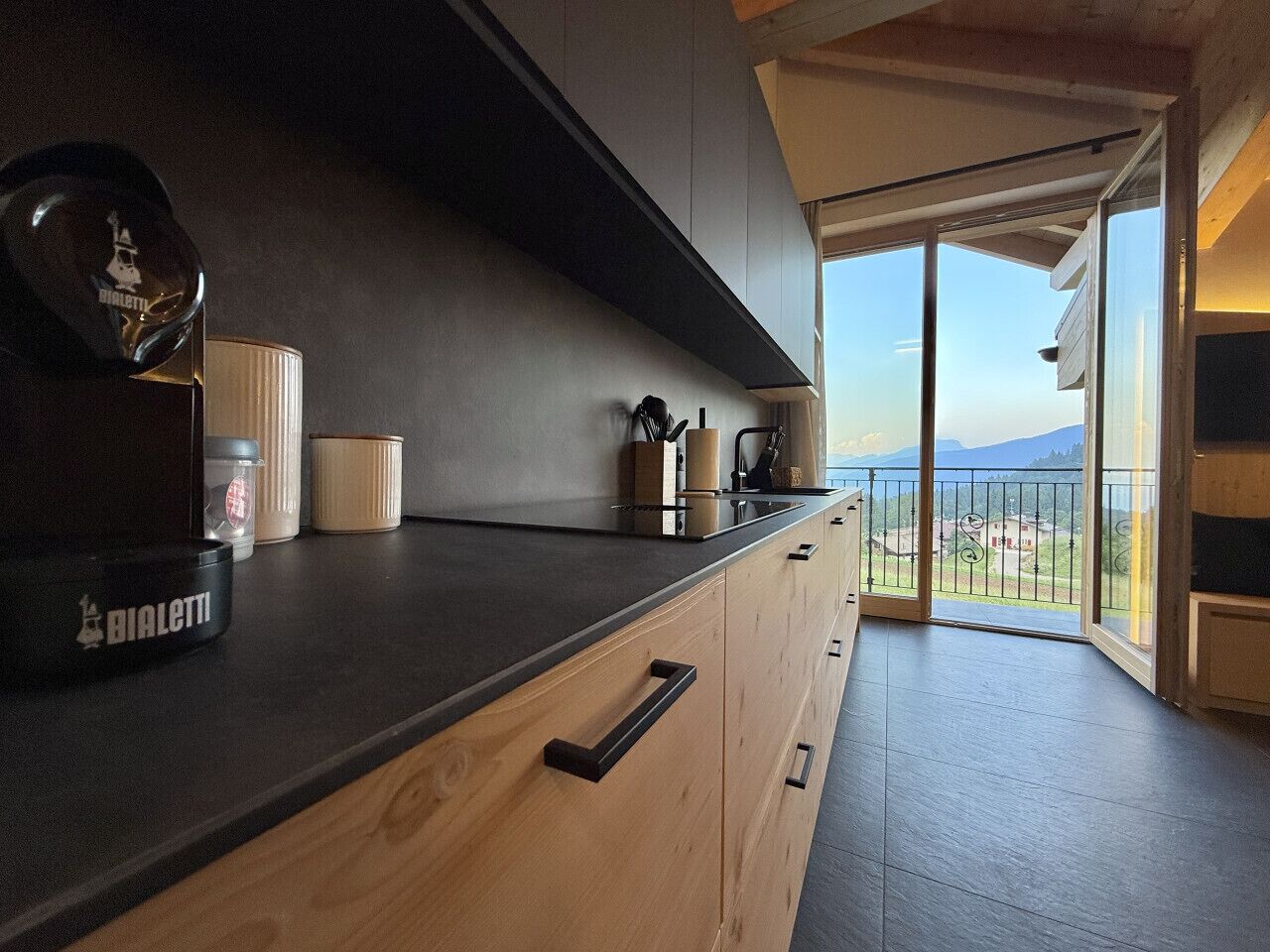 A modern kitchen with a dark countertop, wooden cabinets, and a view of the mountains through a glass door.