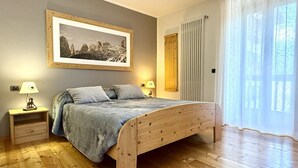 Een slaapkamer met een houten bed, een nachtkastje met een lamp, een ingelijst foto aan de muur, en een raam met gordijnen.