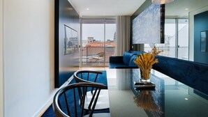 A modern living room with a glass dining table, two chairs, a blue sofa, and a large window offering a city view.