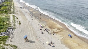 Ein Strand mit einer weiten Sandfläche, einigen Sonnenschirmen und einer Holzpromenade, die zu einem Gebäude führt.