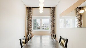 A dining room with a long table, chairs, patterned curtains, and a view of buildings through a window.