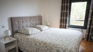 Une chambre à coucher avec une tête de lit capitonnée, un lit avec une literie à motifs, une table de chevet avec une lampe et une fenêtre avec des rideaux.